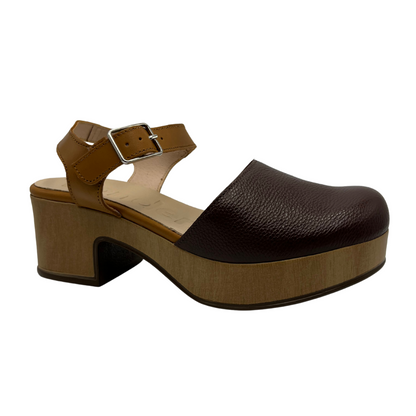 Angled view of dual toned brown leather clog with a thick wood toned outsole and chunky heel. Silver buckle strap and closed toe.