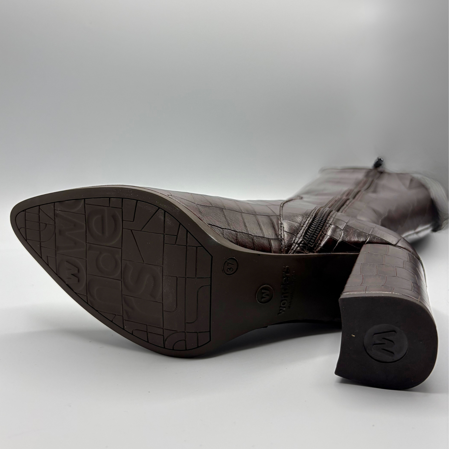 A dark brown crocodile-embossed leather boot shown from the sole and heel end, featuring a pointed toe, side zipper, and a block heel with a monogram 'W' on the heel cap.