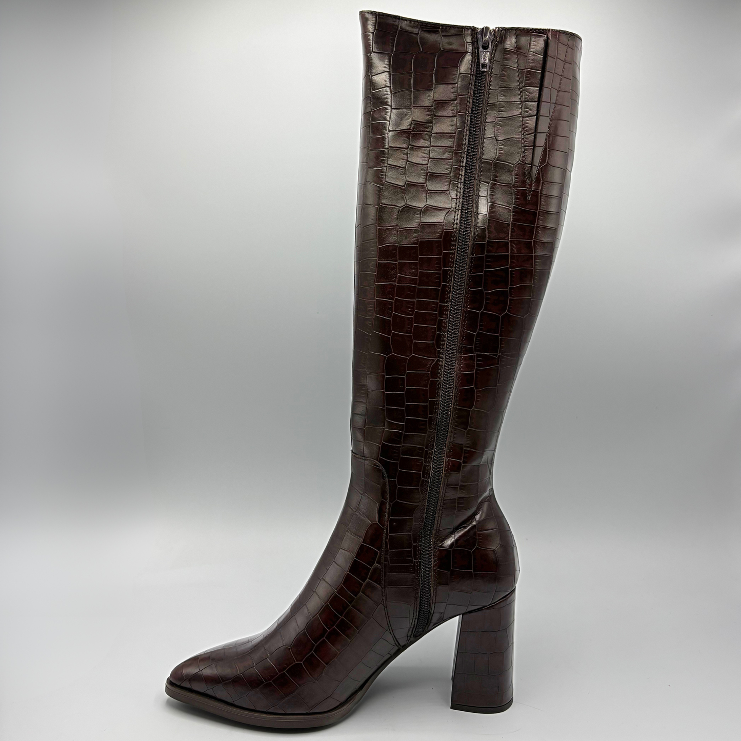 A single knee-high boot in glossy dark brown crocodile-embossed leather, featuring a pointed toe and a chunky block heel, shown against a neutral gray background.