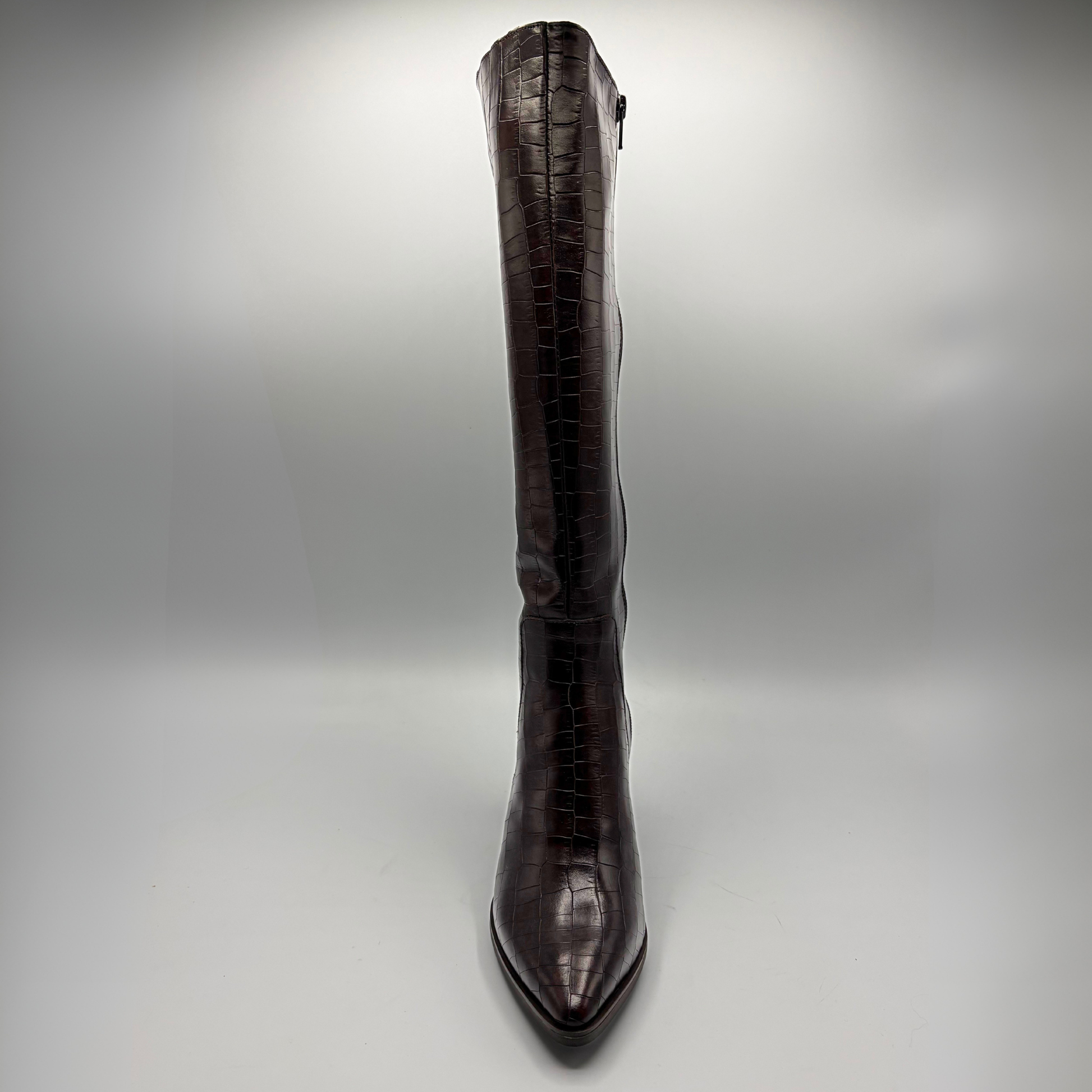 A single knee-high pointed-toe boot with a glossy crocodile-embossed texture in a deep dark brown color, photographed against a neutral gray studio background.
