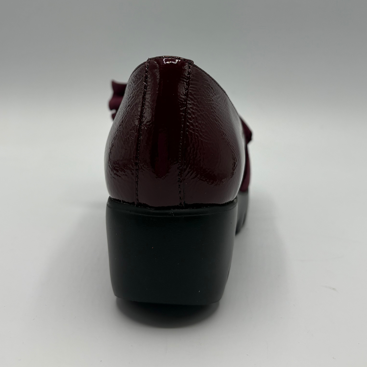 A close-up rear view of a shoe in glossy burgundy patent leather with a chunky black block heel, shown on a neutral gray background.