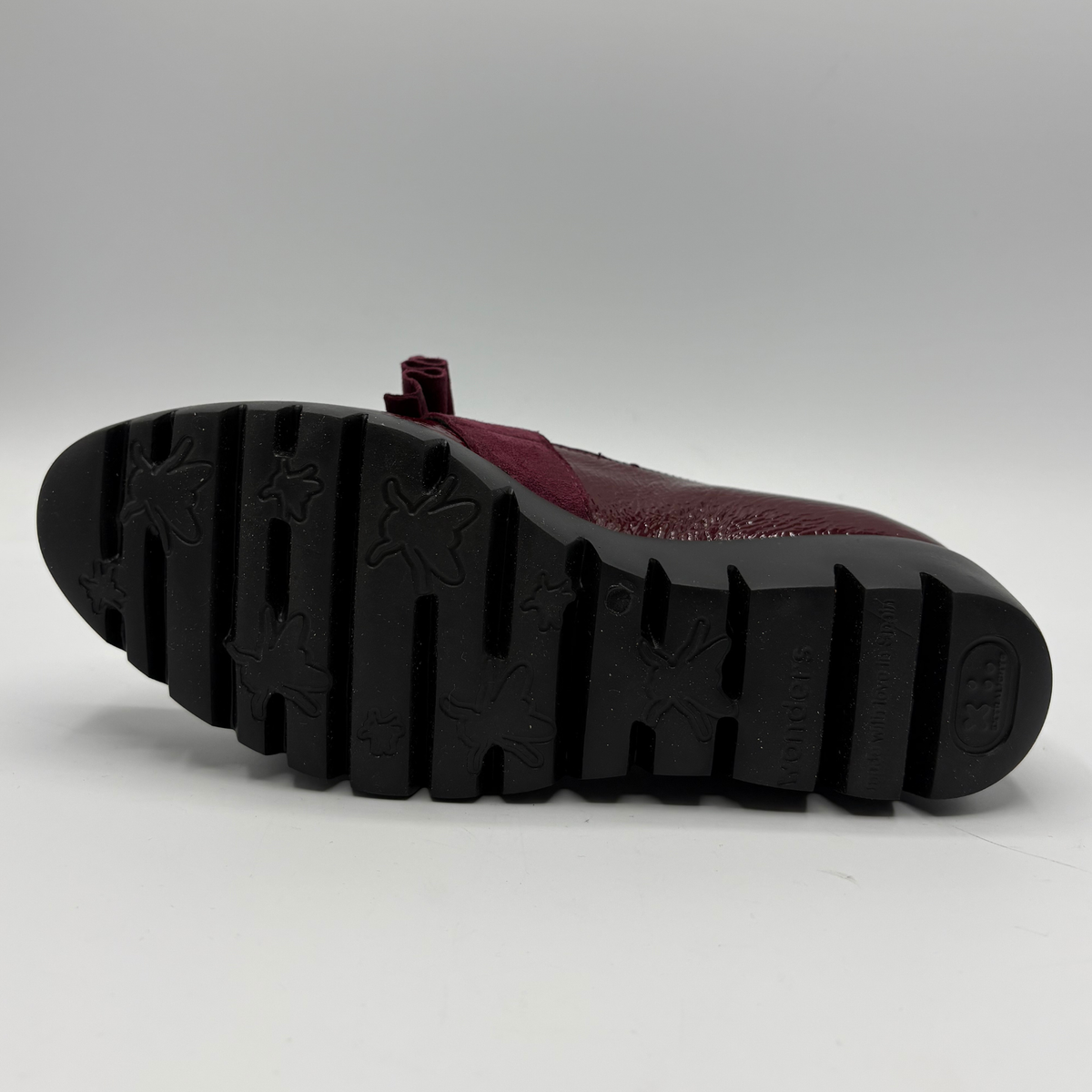 A close-up of a shoe's sole and heel, showing a textured black rubber tread with  a burgundy textured upper.