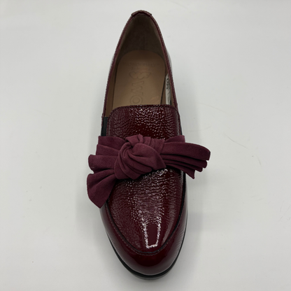 A single women's loafer in deep burgundy, featuring a glossy textured patent leather toe cap, a suede-like bow detail across the vamp, and a black outsole, shown from a top-down angle on a white background.