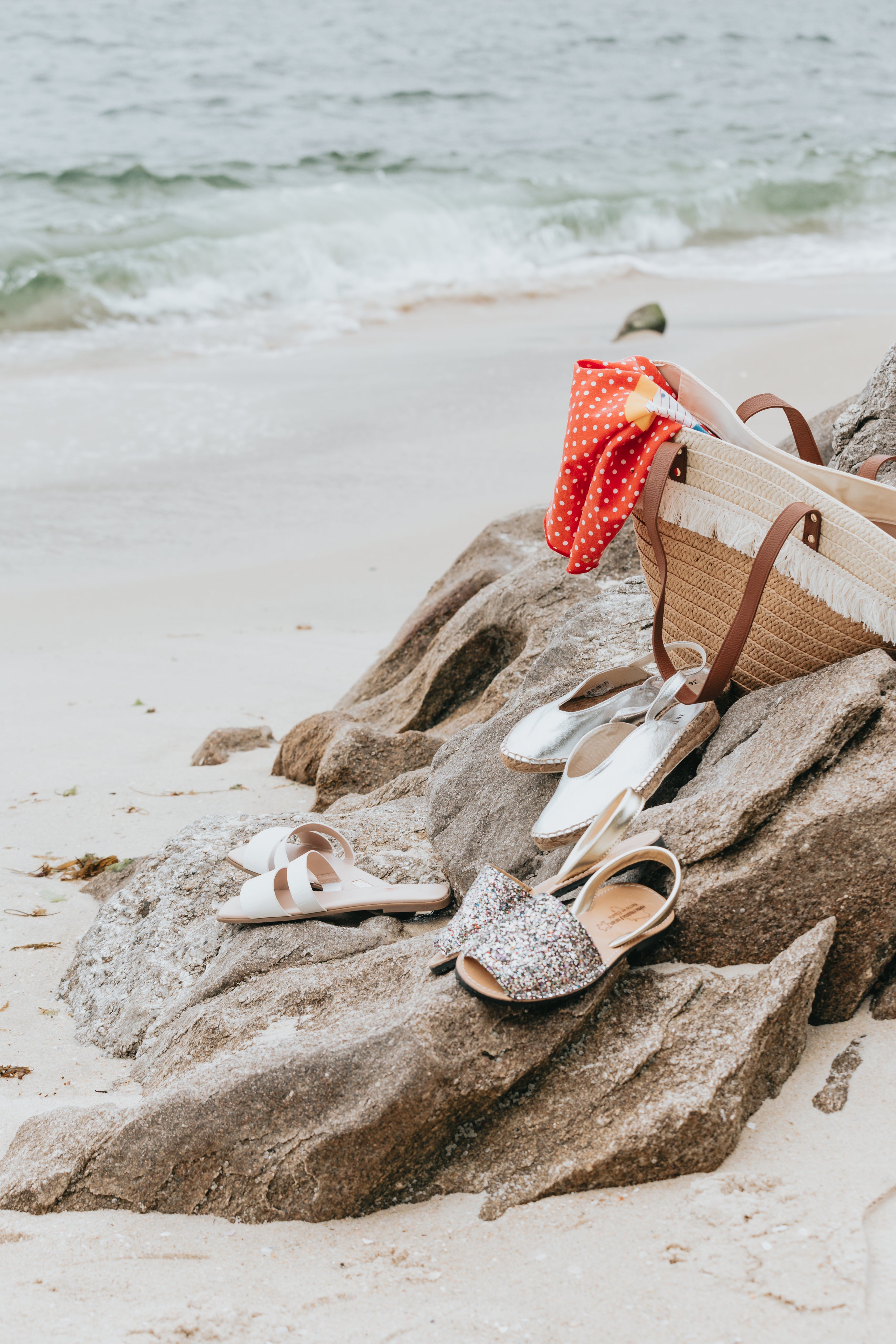 Three pairs of summer shoes sit on top of the rocks on a sandy beach with a wicker beach bag behind them.
