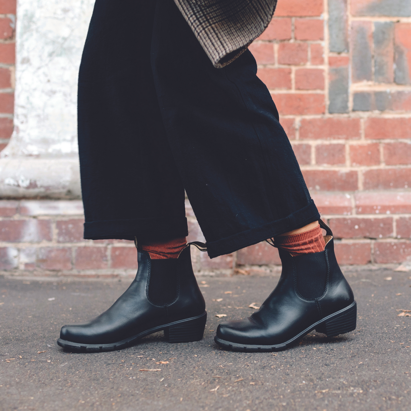 Person wearing black, leather, mid-heeled boot in front of a brick wall