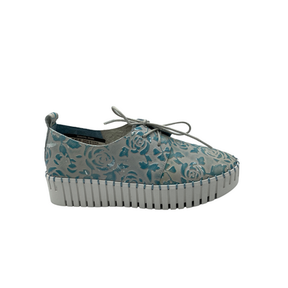 Outside view of sneaker style loafer in a denim blue with embossed florals in the leather.  Laces on front for adjustable fit.  Lined rubber sole