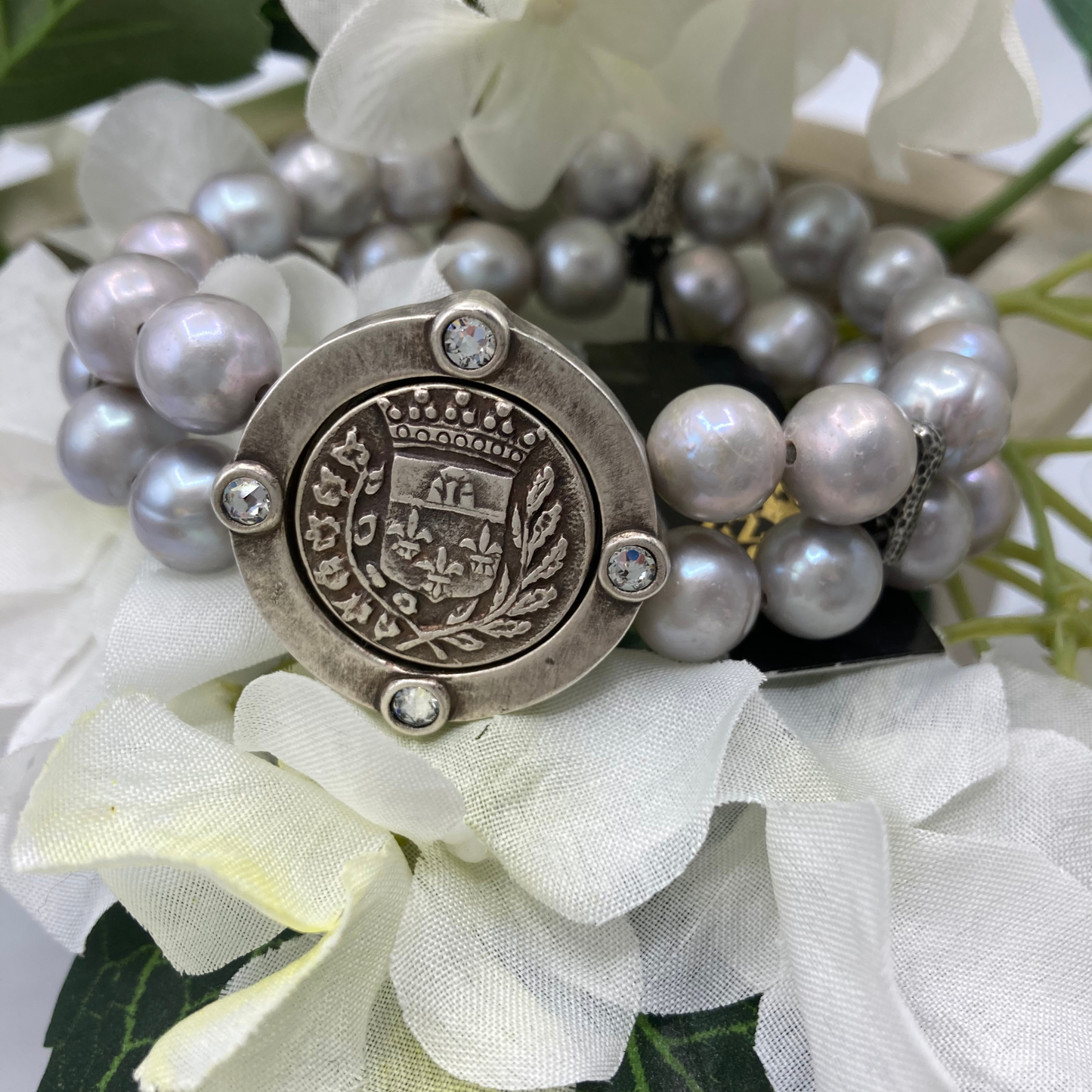 A silver pearl bracelet is pictured featuring a silver coat of arms medallion encrusted with 4 small diamonds on the periphery. 