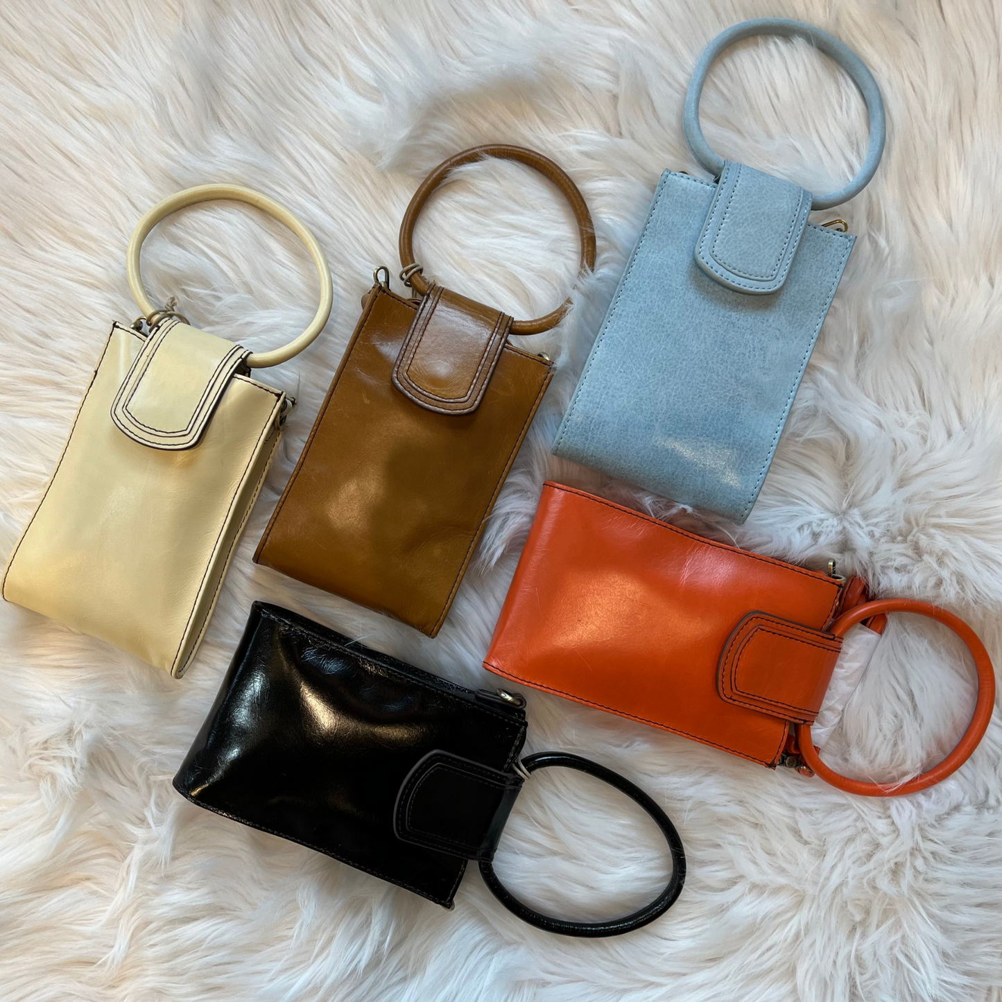 A grouping of 5 Sheila Phone Crossbody bags, 3 across the top and the 2 on the bottom. They are pictured on a white fur rug. There are 5 colours, Butter, Truffle, Blue Topaz, Black and Zinnia. The bags are shown without the optional crossbody strap.