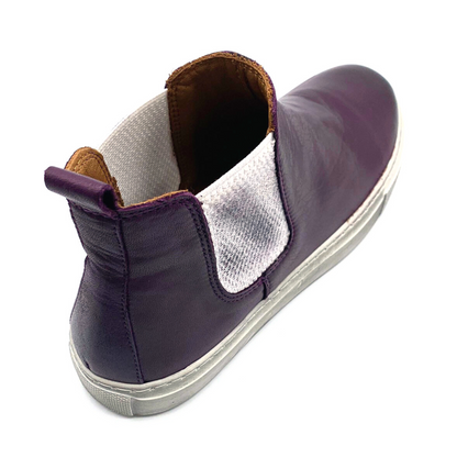 A dark purple shoe is pictured from behind at an angle with white sole, white side elastic, tan inner leather, and a heel pull tab.