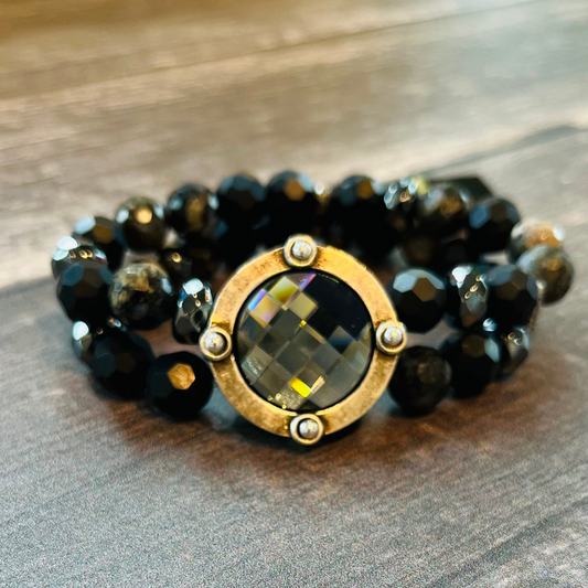 A beautiful handmade bracelet is pictured. It has a larger, circular, black Austrian Crystal centre. The crystal is encased by a gold coloured metal ring with 4 small studs. Twin strands of black beads form the stretch bracelet and attach to the centre crystal.