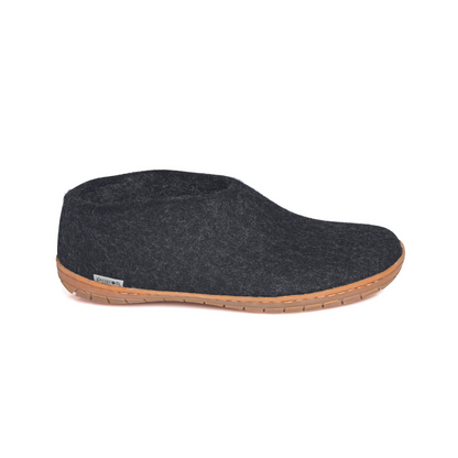 A dark grey slipper is pictured in profile, showing the high back of the heel. The bottom lined in caramel rubber has a small tag with the brand name attached in the seam where the leather meets the felted wool.