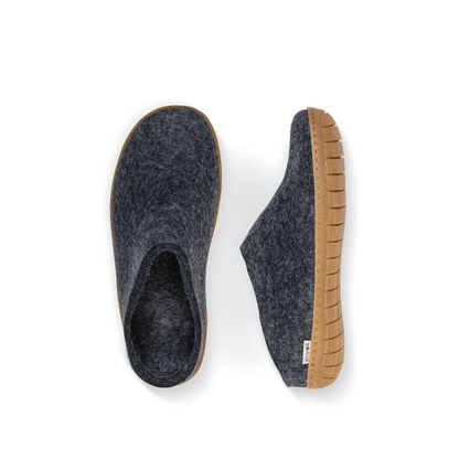 An overhead view of two denim-blue felted wool slippers. One is pictured straight on, showing the top face of the slipper, while the other lays on its side, showing the profile with caramel rubber sole with a row of stitching.