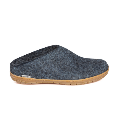 A denim-blue slipper is pictured in profile, showing the low back of the heel. The bottom lined in caramel coloured rubber has a small tag with the brand name attached in the seam where the rubber meets the felted wool.