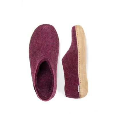 An overhead view of two dark-mauve felted wool tall-back slippers. One is pictured straight on, showing the top face of the slipper, while the other lays on its side, showing the profile with tan leather sole with a row of stitching.