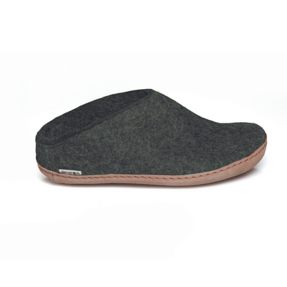 A dark green slipper is pictured in profile, showing the low back of the heel. The bottom lined in tan leather has a small tag with the brand name attached in the seam where the leather meets the felted wool.